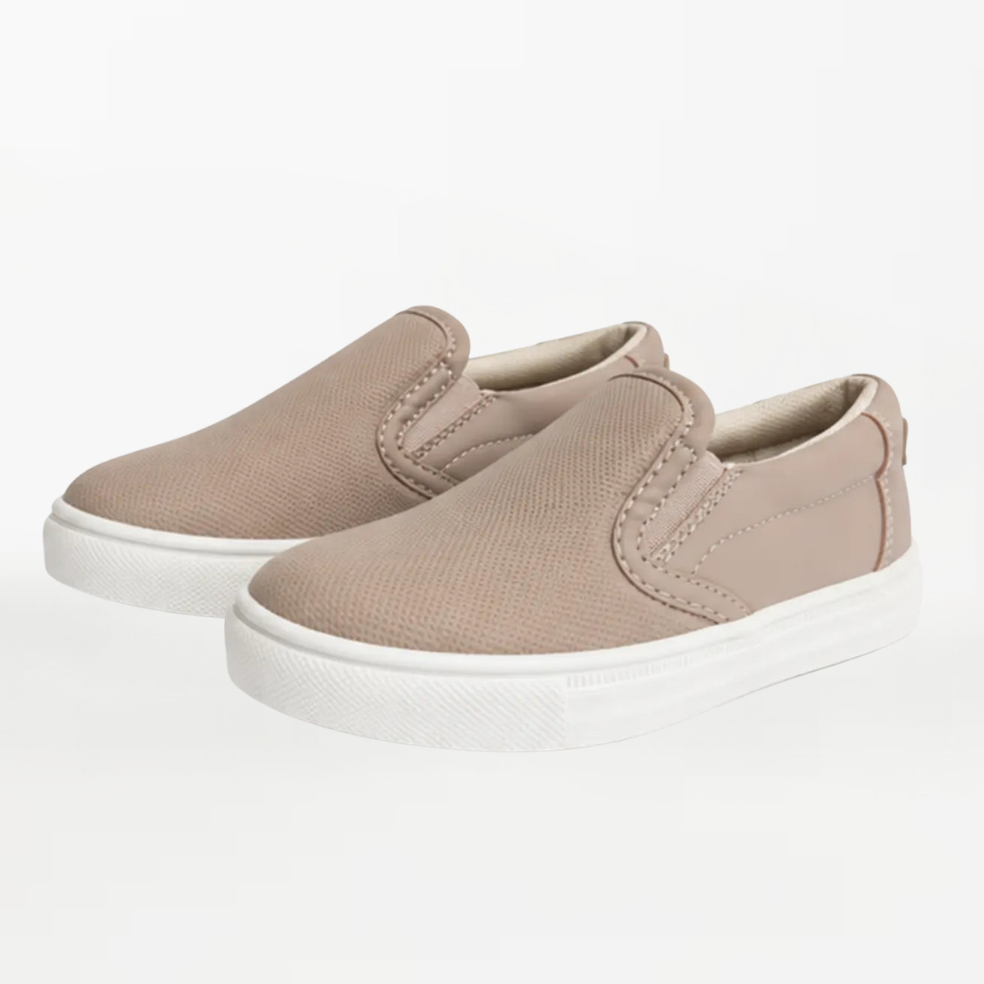 A pair of toddler slip-on sneakers in a light taupe-beige canvas with white rubber soles, photographed on a seamless white studio background.