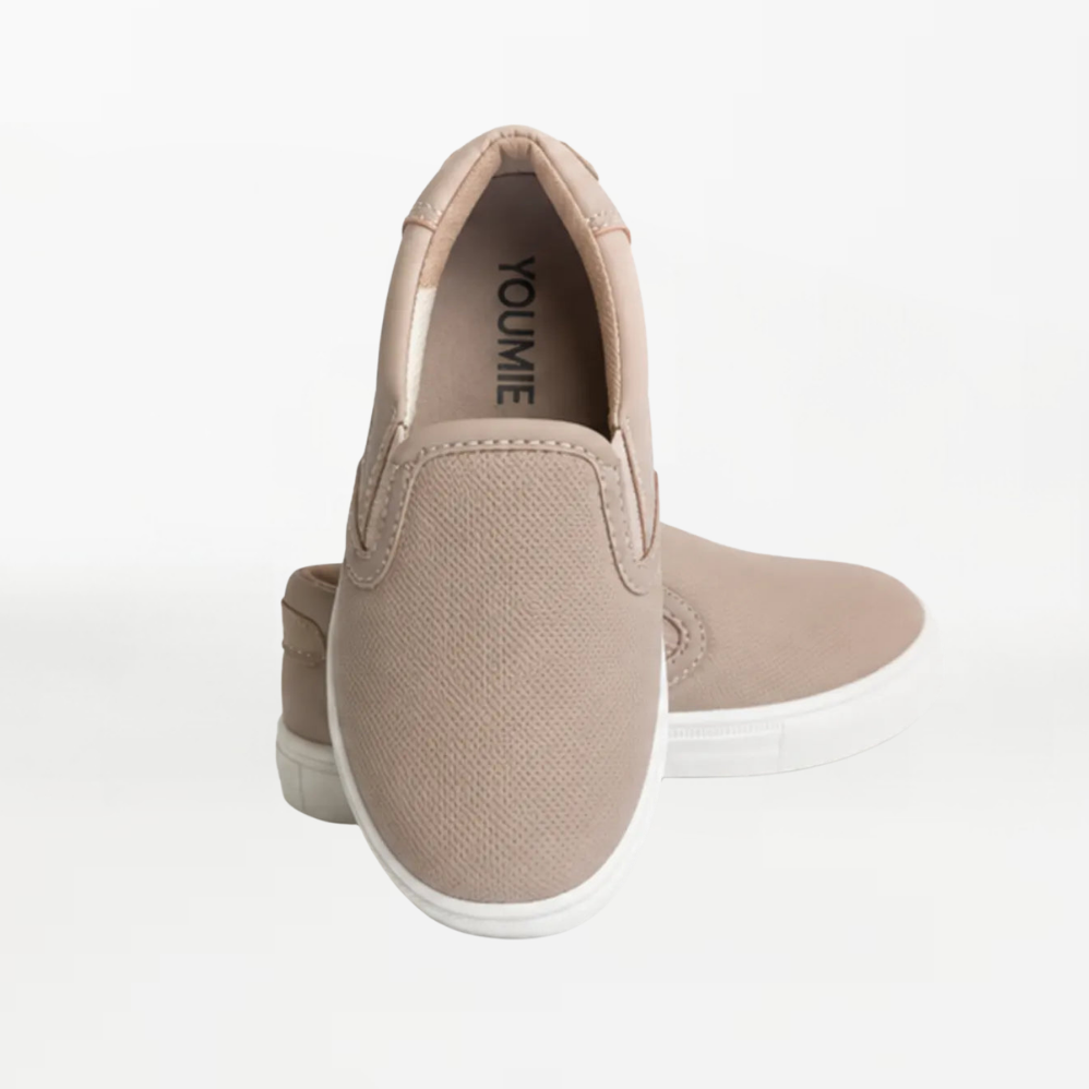 A pair of slip-on canvas sneakers in a light taupe-beige color with white soles, shown from a rear three-quarter angle on a clean white background.