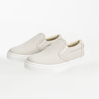 A pair of slip-on canvas sneakers in a light beige color with white soles, photographed on a seamless white background.