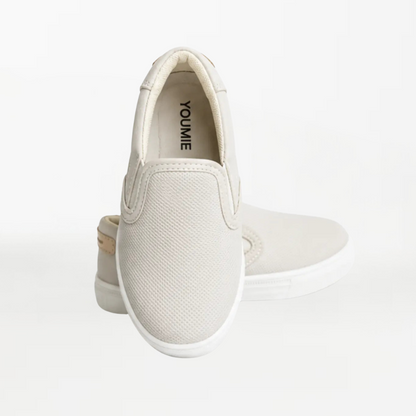 A pair of slip-on canvas sneakers in a light cream color, shown from a rear three-quarter angle on a seamless white background.